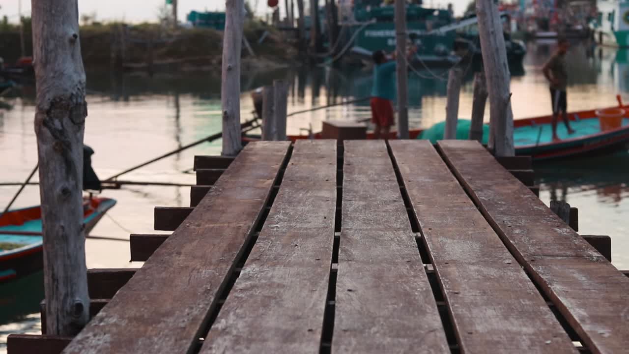 Focus is on the handmade wooden bridge whilst a local thai fishing boat slowly flows past out of focus in the background during the sunset. The water has a golden glow to it.