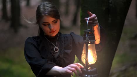 Beautiful-girl-with-long-hair-and-a-vintage-dress-adjusts-the-brightness-of-a-kerosene-lamp-to-illuminate-herself-in-a-dense-dark-forest