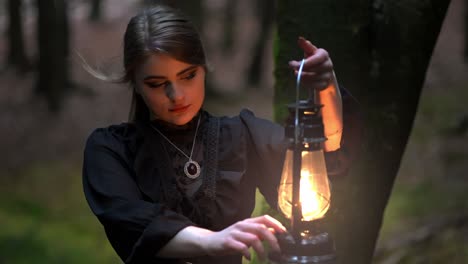 hermosa chica con cabello largo y un vestido vintage ajusta el brillo de una lámpara de queroseno para iluminarse en un denso bosque oscuro