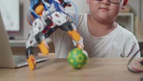 primer plano de un chico asiático con una mano de cyborg tratando de sostener una pelota pequeña mientras trabaja en una computadora portátil en casa