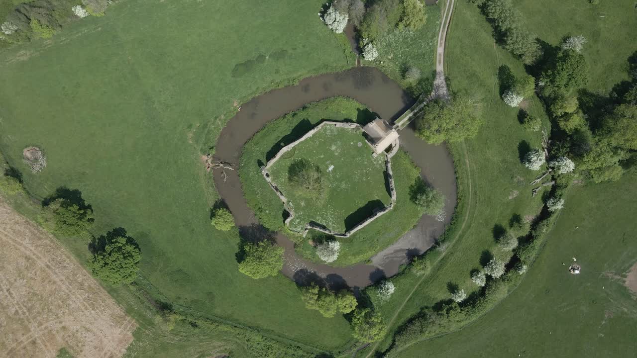 Aerial view of Stogursey Castle, a medieval castle in Somerset, England. Most of the site is in ruins, with a thatched gatehouse. Drone descending and rotating to the right over the ruins and moat