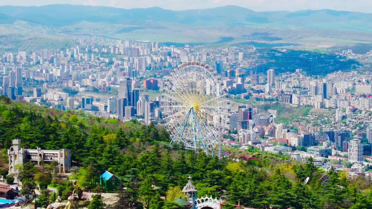 Veduta aerea con drone di Tbilisi dal Monte Mtatsminda, con ruota panoramica, skyline della città, fiume e ampi paesaggi urbani.