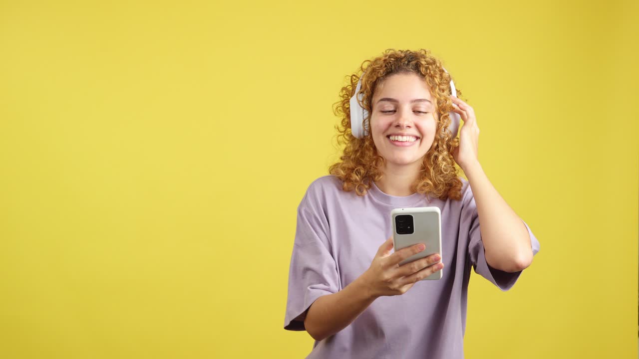 Young Woman with Curly Hair Enjoying Music with Headphones and Smartphone on Yellow Background