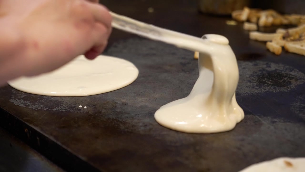 Ladle pours smooth pancake batter onto a hot griddle, spreading to form two perfect rounds in a bustling restaurant kitchen. Warm lighting highlights the delicious breakfast preparation. Handheld 4K