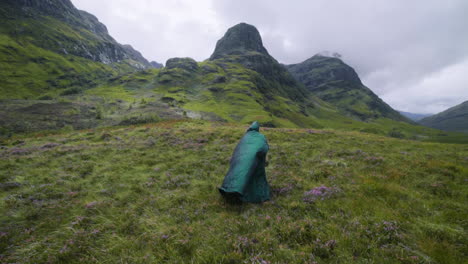 cloaked figure walks up green hill to an outcropping of rocks in the scottish highlands