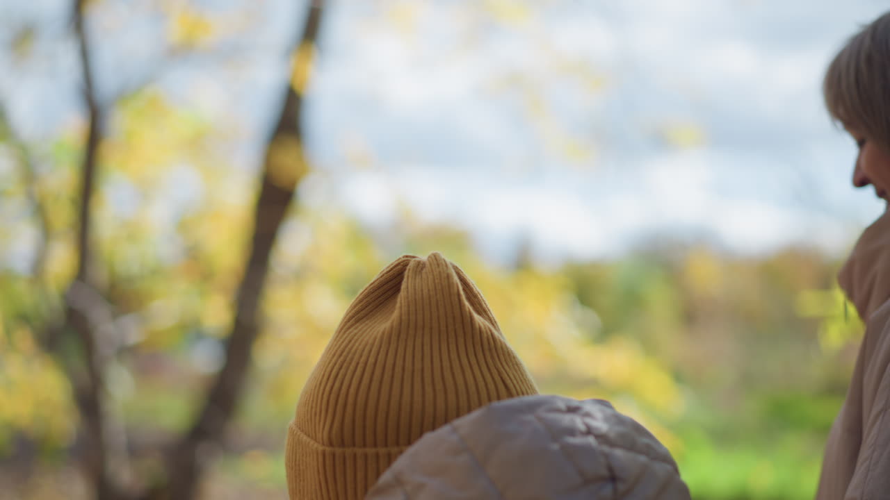 Close up rear view of kid wearing hooded jacket and beanie walking with aunt through sunlit autumn forest, engaging warmly as golden leaves blur gently in background breeze