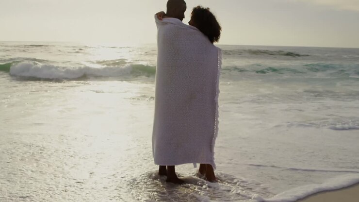 Vista laterale di coppia afroamericana avvolta in una coperta sulla spiaggia 4K
