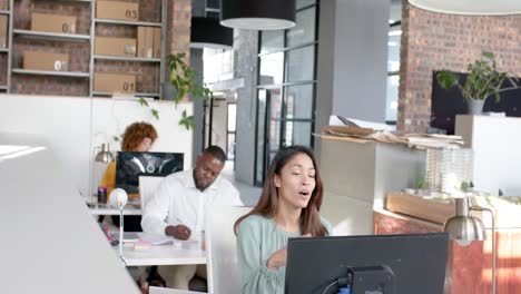 Mujer-De-Negocios-Informal-Que-Tiene-Videollamada-Con-Computadora-Portátil-En-Una-Oficina-Ocupada-Con-Colegas-Diversos,-Cámara-Lenta