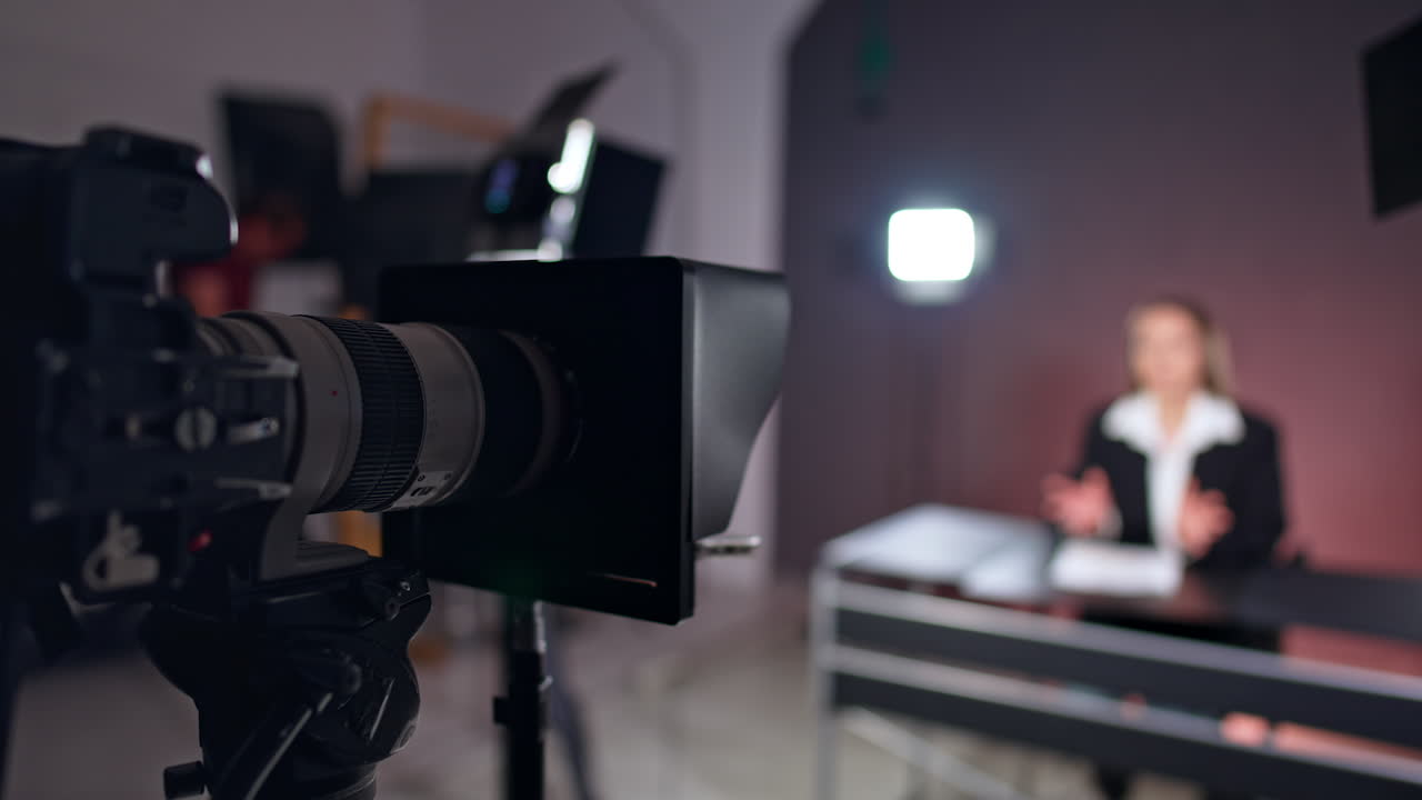 Professional camera with a teleprompter. Female blogger sitting at desk at blurred backdrop.