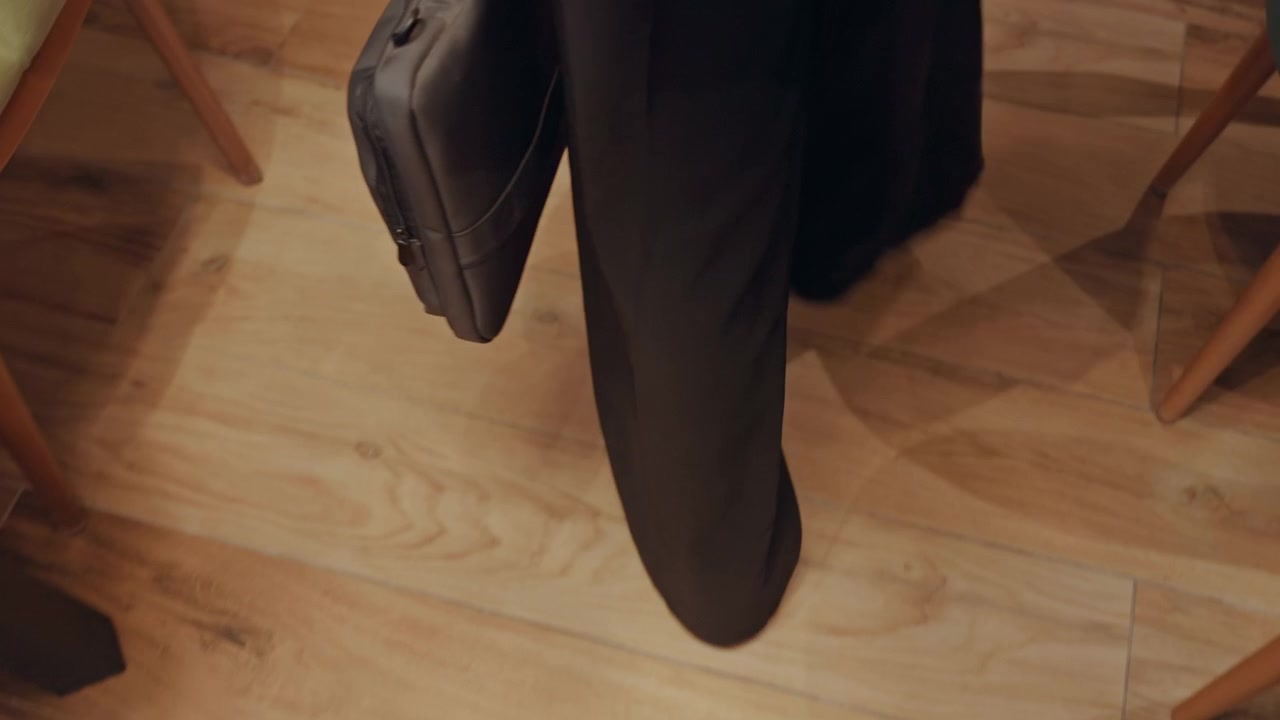 Gambe che camminano su un pavimento in legno, dirigente d'azienda in abito che porta una ventiquattrore, passo misurato attraverso una hall tranquilla, scarpe lucide, contrasto d'ombra, avvicinamento a una riunione interna, texture dettagliata del pavimento