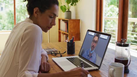 mujer afroamericana tomando notas mientras tiene una videollamada en una computadora portátil en casa