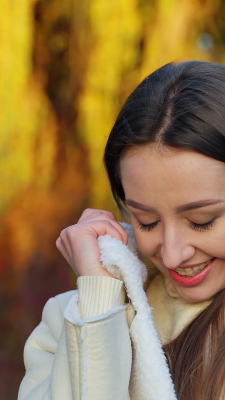 Happy brunette woman enjoying her time in the nature. Smiling lady wrapping herself up in jacket outdoors on autumn day. Blurred backdrop. Vertical video