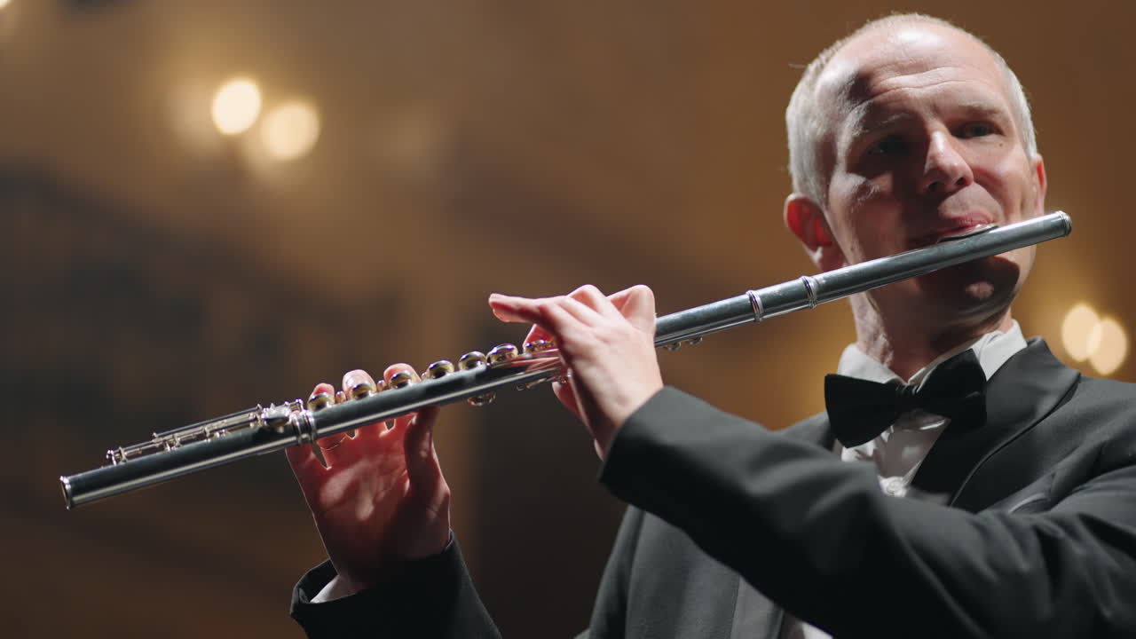 회색 머리카락의 플루트 연주자는 필하모닉 홀에서 음악을 연주하고 있습니다. 심포니 오케스트라의 리허설이나 콘서트입니다.