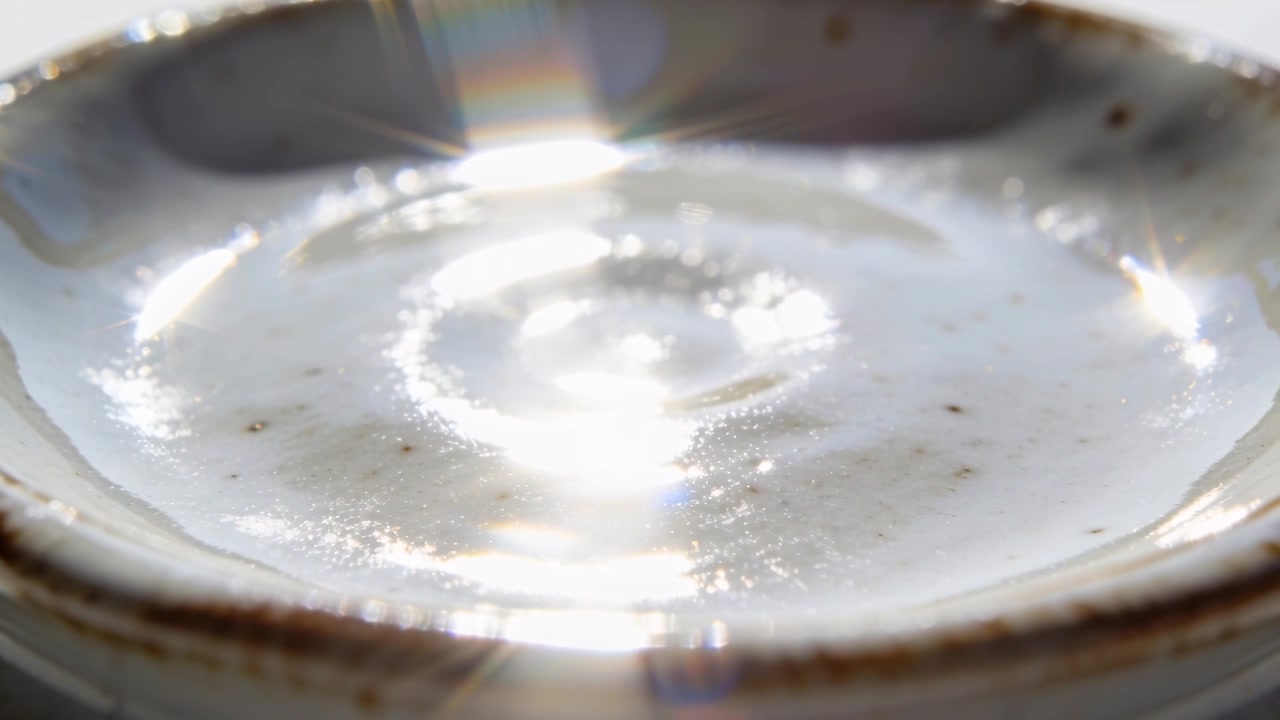 テーブルの上の浅い釉薬のかかったボウルにカメラがズームインし、白い光沢のある液体とスペキュラーフレアが現れる。