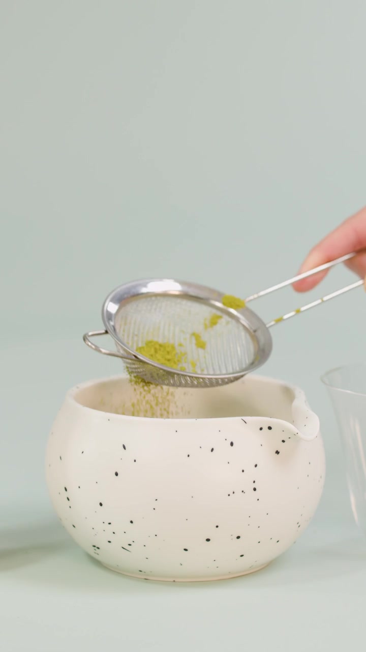 Mano tamiza polvo de matcha verde a través de un colador de malla en un cuenco de cerámica moteado, con iluminación suave de estudio.