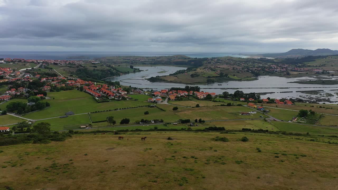 vuelo con un dron en la desembocadura de un río a la marcon un giro de la cámara vemos una colina con caballos pastando y vemos una ciudad con prados verdes en una mañana nublada en cantabria españa