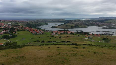 Vuelo-Con-Dron-En-La-Desembocadura-De-Un-Río-Hasta-El-Marcon-Un-Giro-De-Cámara-Vemos-Un-Cerro-Con-Caballos-Pastando-Y-Vemos-Un-Pueblo-Con-Prados-Verdes-En-Una-Mañana-Nublada-En-Cantabria-España