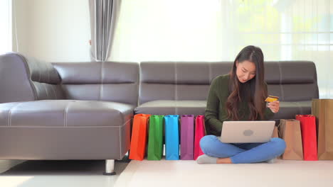 While-sitting-on-the-floor,-a-young-woman-with-a-credit-card-in-one-hand-puts-her-numbers-into-a-website-on-her-laptop