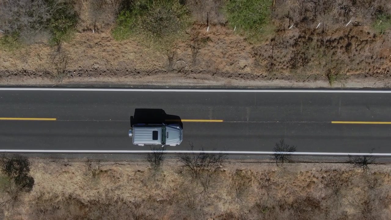 Vista aerea con drone che mostra un'auto che viaggia lungo un'autostrada asfaltata attraverso un paesaggio rurale arido, con alberi sparsi, vegetazione marrone, segnaletica stradale gialla e terreno aperto intorno alla strada
