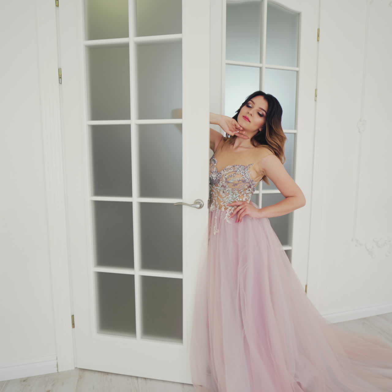Beautiful bride in wedding dress is standing near the door in the light room. Gorgeous model in luxury dress is posing in studio.