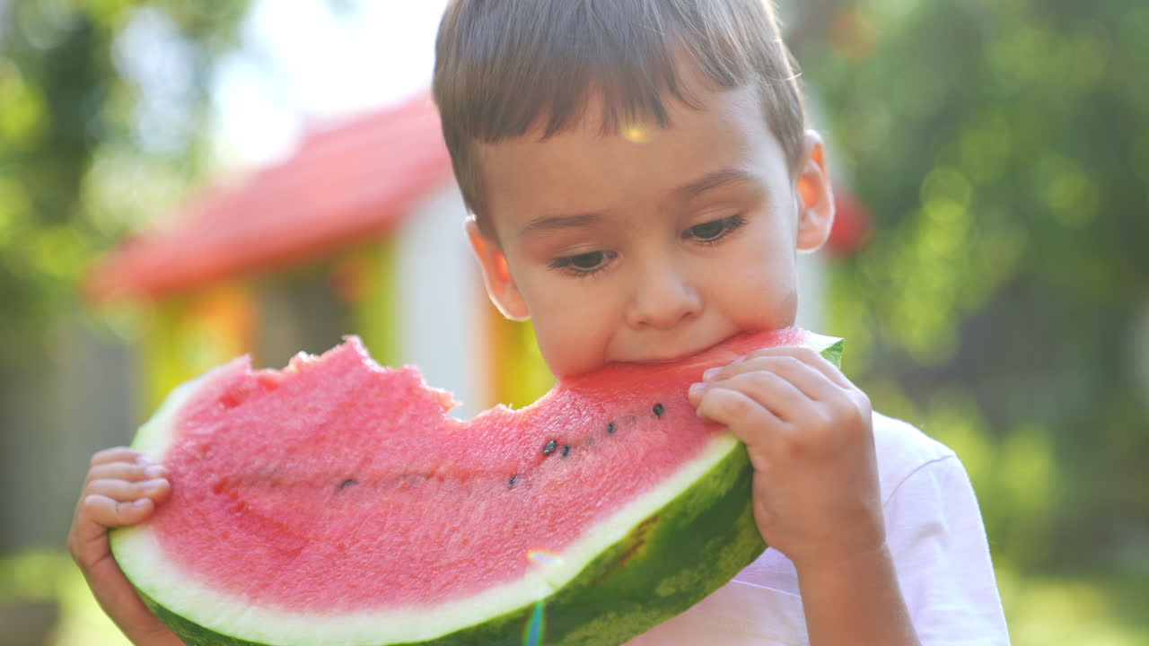 Little boy eating a fresh ripe watermelon outdoors. Fruit juice runs by the kid's chin. Close up. Blurred backdrop.