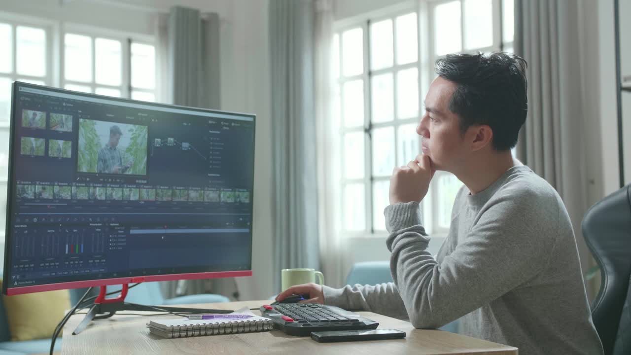 Asian Video Editor Man In Long Sleeved T-Shirt Thinking While Using Desktop Computer For Editing Video At Home.