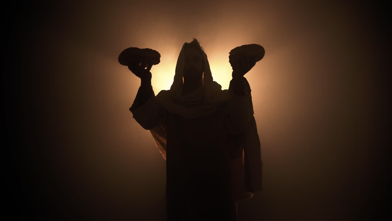 Silhouette of Jesus Christ in white hooded robe holding bread as he stands in glowing, bright light praying. Biblical, Christianity visual for Easter, last supper, communion.