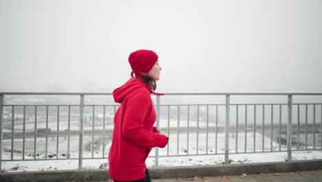 vista lateral de una mujer de negocios corriendo con gorra roja y chaqueta a lo largo de un camino nevado cerca de una barandilla de hierro, pareciendo agotada, con el paisaje urbano y el puente en una atmósfera de invierno brumosa