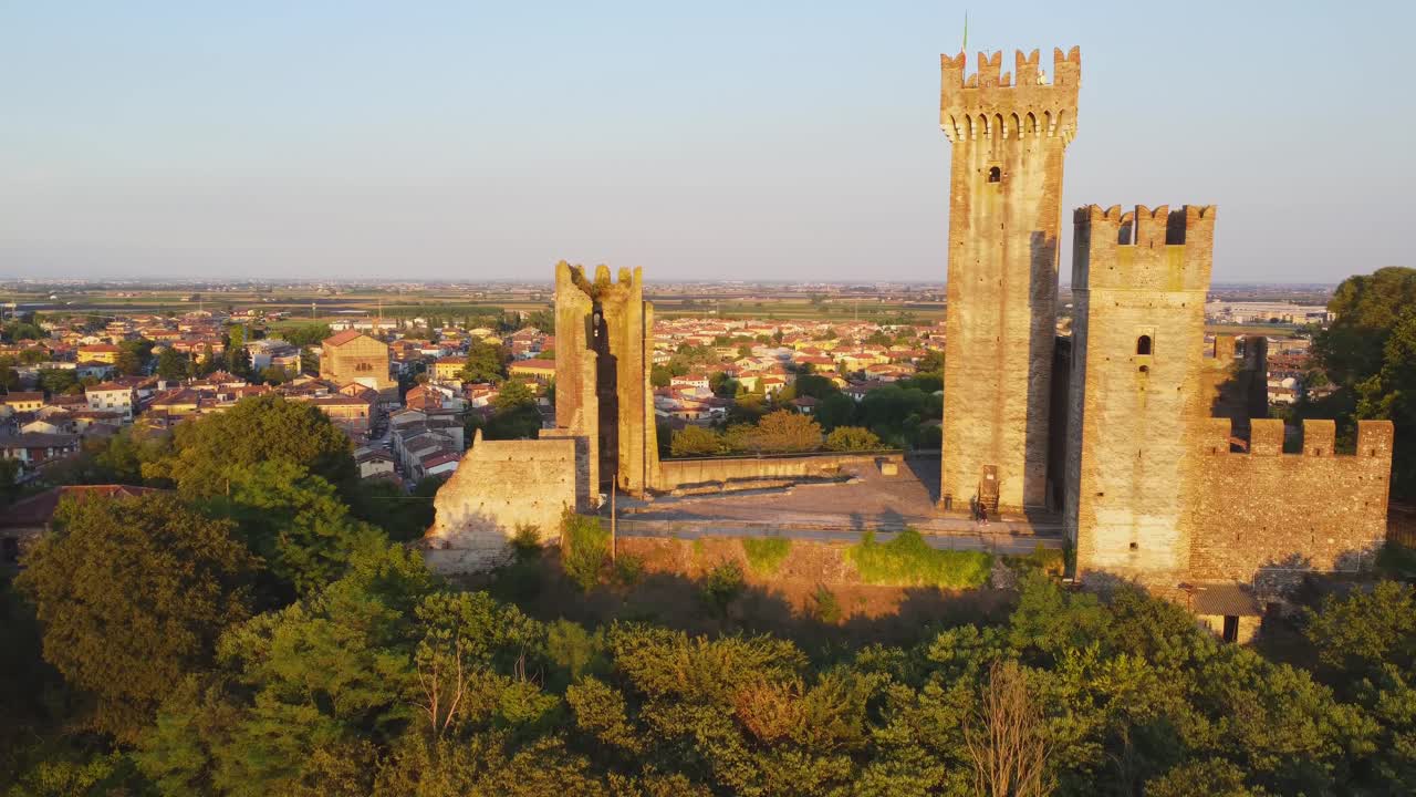 A golden hour drone view of Castello Scaligero in Valeggio sul Mincio. The medieval fortress rises above the trees, overlooking the Veneto plains in a breathtaking warm evening light.