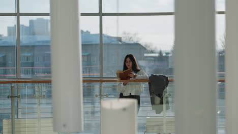 focused young woman stands reading novel near indoor glass railing, natural daylight casting soft glow through large windows, with urban background of residential rooftops