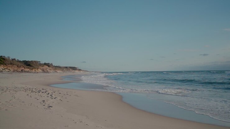 Idyllischer Sandstrand mit sanften Wellen und einer fernen Steilküste unter einem klaren blauen Himmel