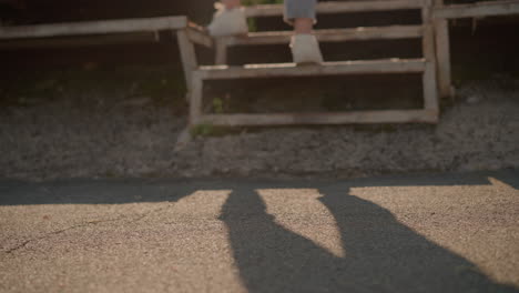 blurred close-up leg view of person in white sneakers and jeans walking toward rusty staircase and climbs it with individual shadow cast on ground in soft sunlight