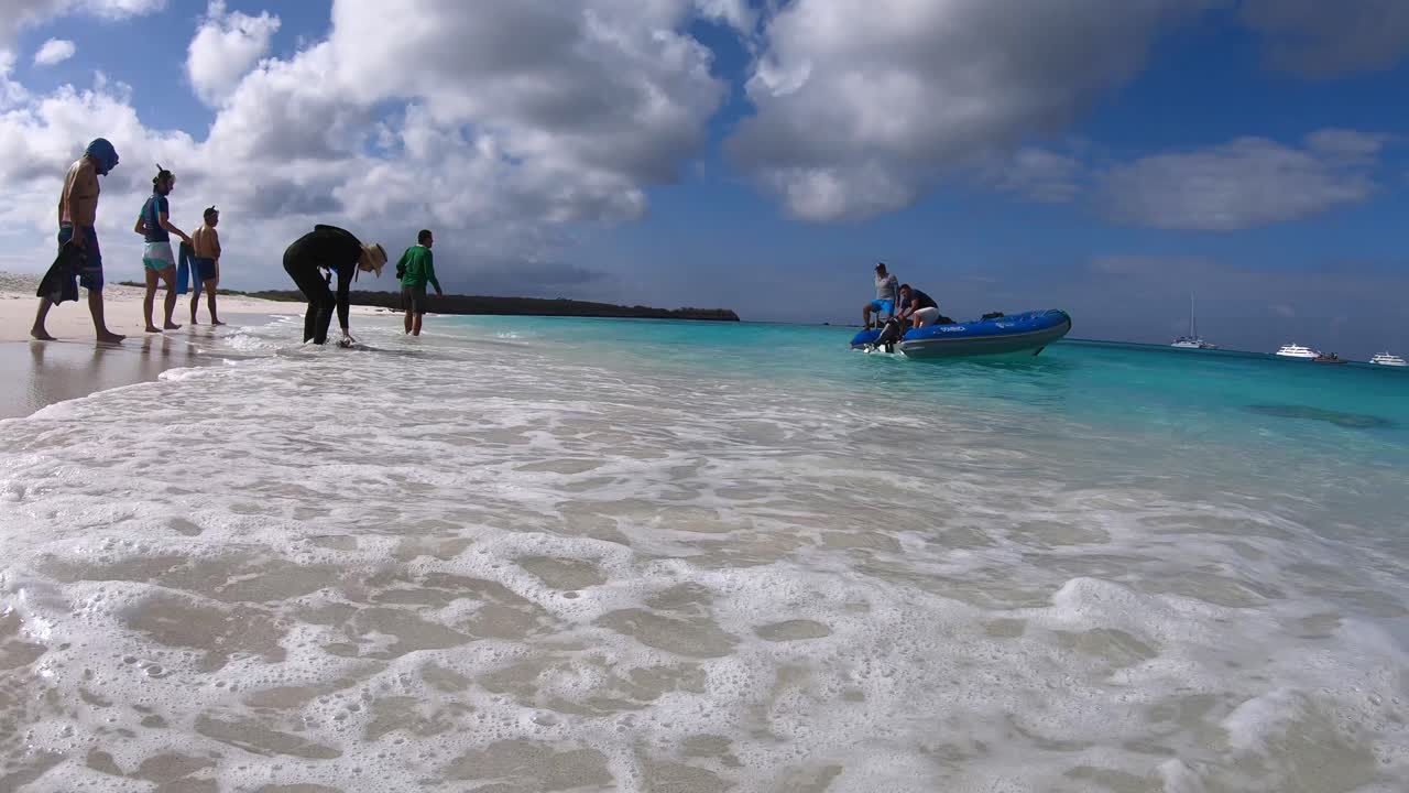 A boat is approaching the shore of a white sand and turquoise water beach of the Galapagos islands while tourists are coming close to the boat to board it
