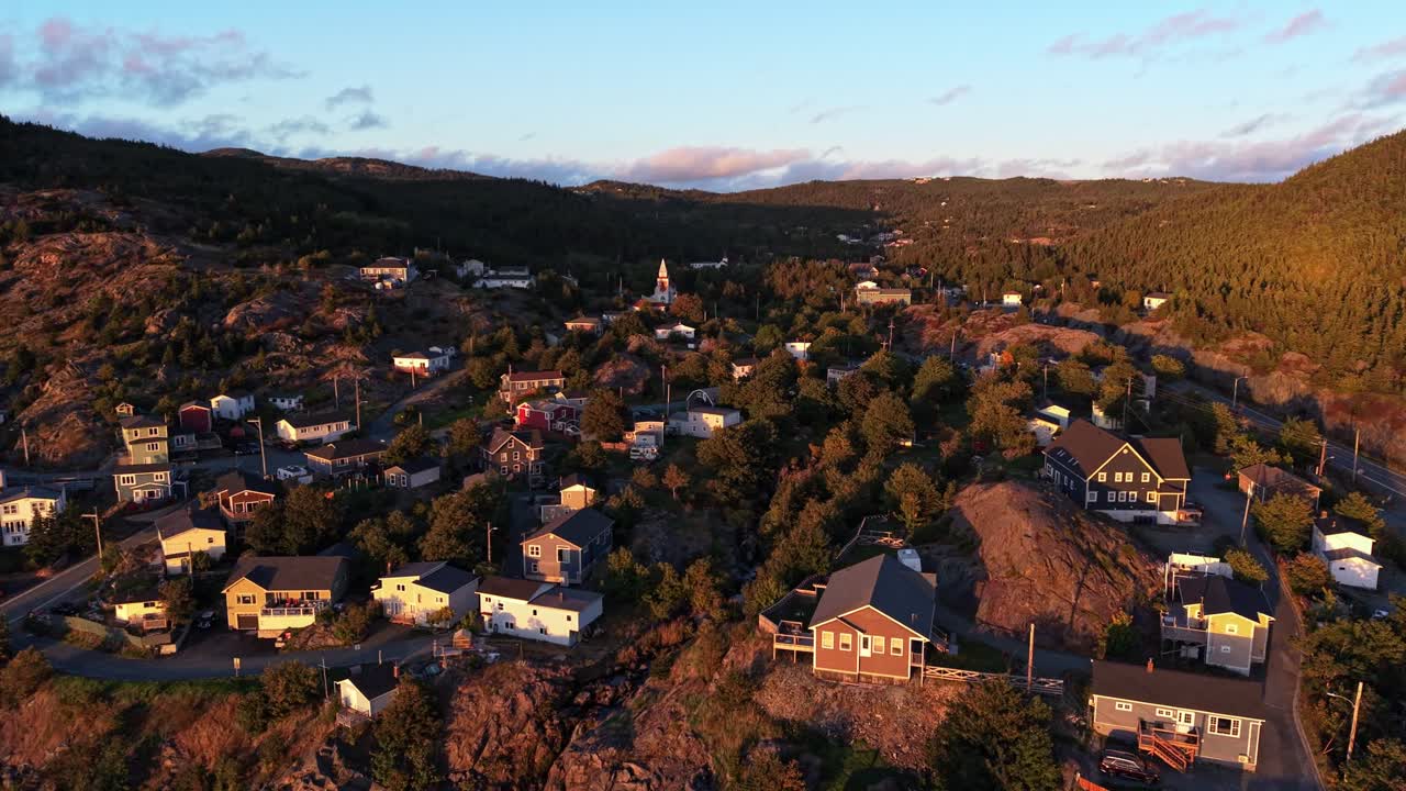 Drone footage reveals Portugal Cove village nestled in a valley with rolling hills and a warm sunset glow, showcasing houses, trees and rugged landscape in Newfoundland