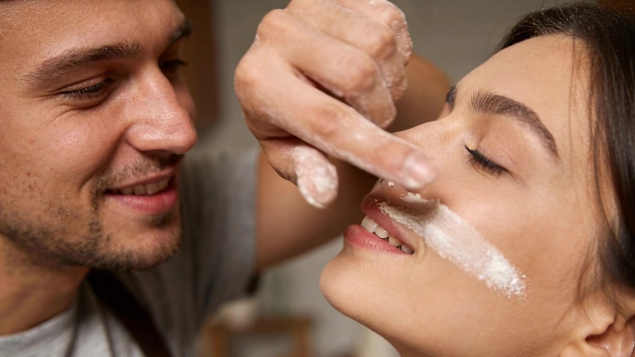 Raising man pressing flour-coated finger on woman's lip during baking in home kitchen, with apron