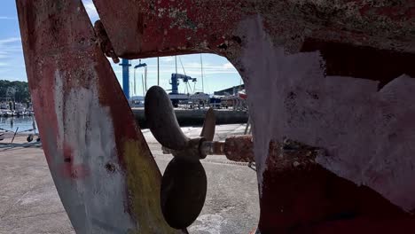 The-rusty-engine-and-rudder-propeller-of-the-old-fishing-boat-in-the-shipyard-for-restoration-with-the-harbor-cranes-in-the-background,-close-up-shot-traveling-backwards