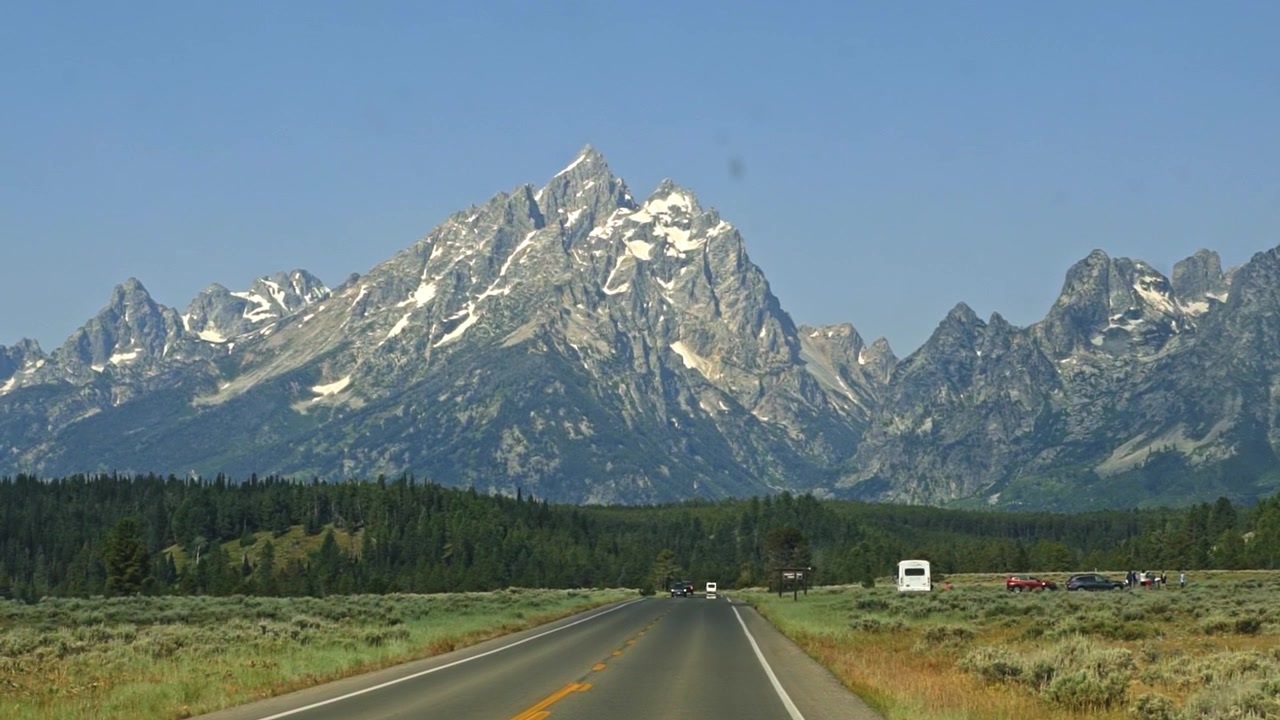 Die Fahrt durch den Grand Teton Nationalpark offenbart die aufragende Teton-Gebirgskette jenseits einer bewaldeten Landschaft und Parkstraße, mit Fahrzeugen darunter, während sich die Kamera sanft durch Wyoming vorwärtsbewegt