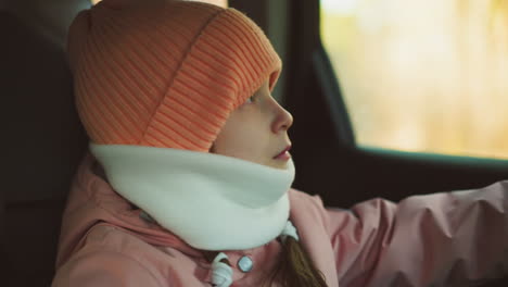 a contemplative moment as a little girl in a pink cap, jacket, and white winter scarf sits in a car, gently placing her hand on the window