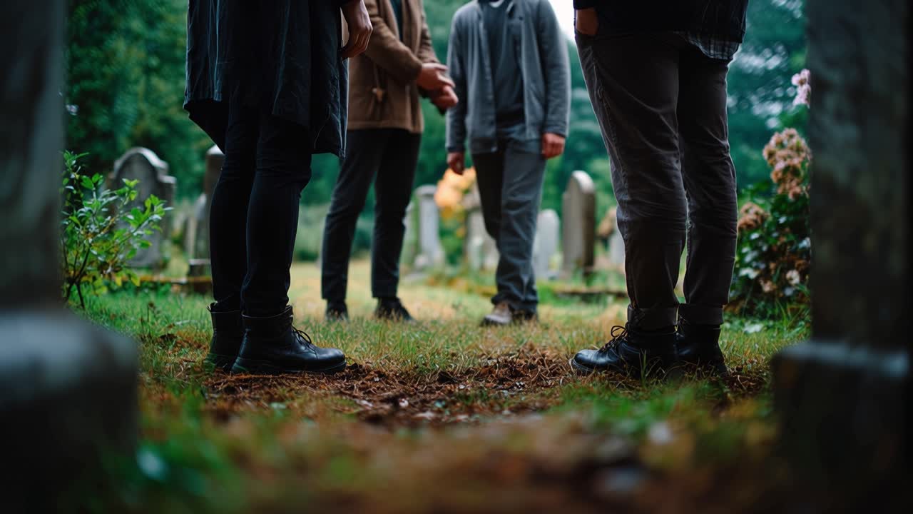 Mourners walk through a quiet, mossy cemetery. Grassy path curves between ancient headstones. One person holds another's hand, showing support. Sadness hangs in the air as they move slowly forward