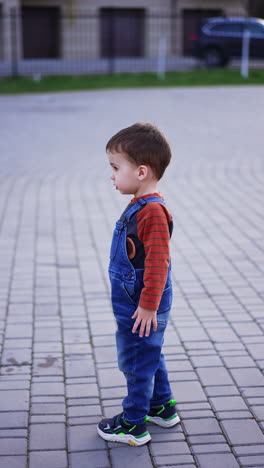 dark-haired caucasian baby boy stands outdoors wearing a shirt and jeans romper. kid turns to camera showing three fingers and runs away. vertical video.