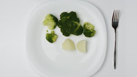 topdown view boiled broccoli and potato plate, hand adding fork on white table