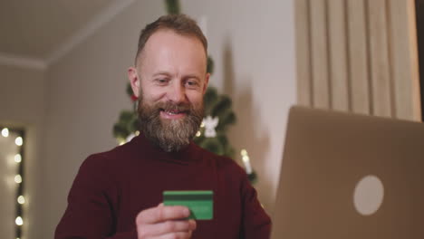 vista inferior de un hombre de cabello rojo que compra en línea con una tarjeta de crédito usando una computadora portátil sentada en una mesa en una habitación decorada con un árbol de navidad 2