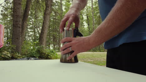 man at forested campsite screws top of moka pot to its base, carries it away