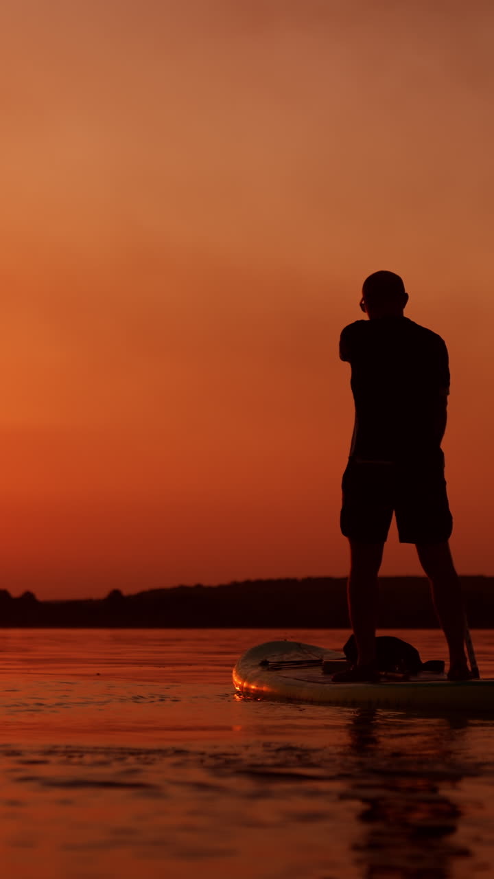Footage of a man sup boarding by the river at sunset. Sportsman rows slowly using a long paddle. Low angle view. Vertical video