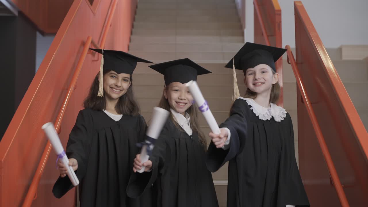 졸업식에서 졸업장을 보여주고 카메라를 바라보는 모자와 가운을 입은 행복한 취학 전 여학생 3명