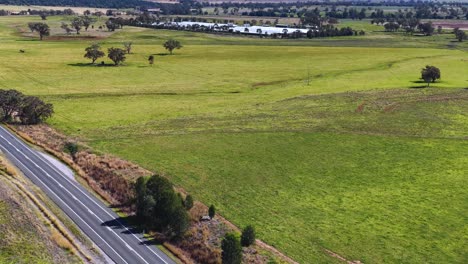 A vehicle travels along a winding rural road bordered by green fields and scattered trees under bright daylight, captured in a smooth aerial tracking shot