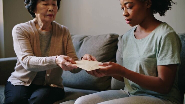 Extending elder in cardigan handing envelope to woman in tee at home on sofa for family