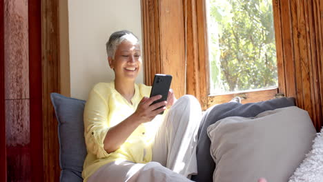 Feliz-Mujer-Birracial-Mayor-Sentada-En-El-Sofá-Y-Usando-Un-Teléfono-Inteligente-En-Casa,-Cámara-Lenta