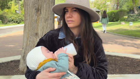 close-up of a young mother feeding her baby with a bottle while sitting on a bench in a tranquil park. natural outdoor setting with emotional connection