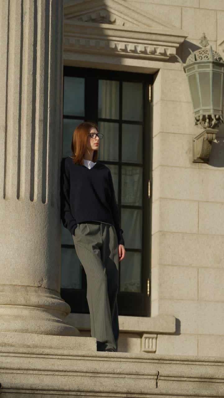 Long lens parallax shot of stylish young woman in glasses and loose trousers posing against massive stone column on Seokjojeon palace steps at golden hour